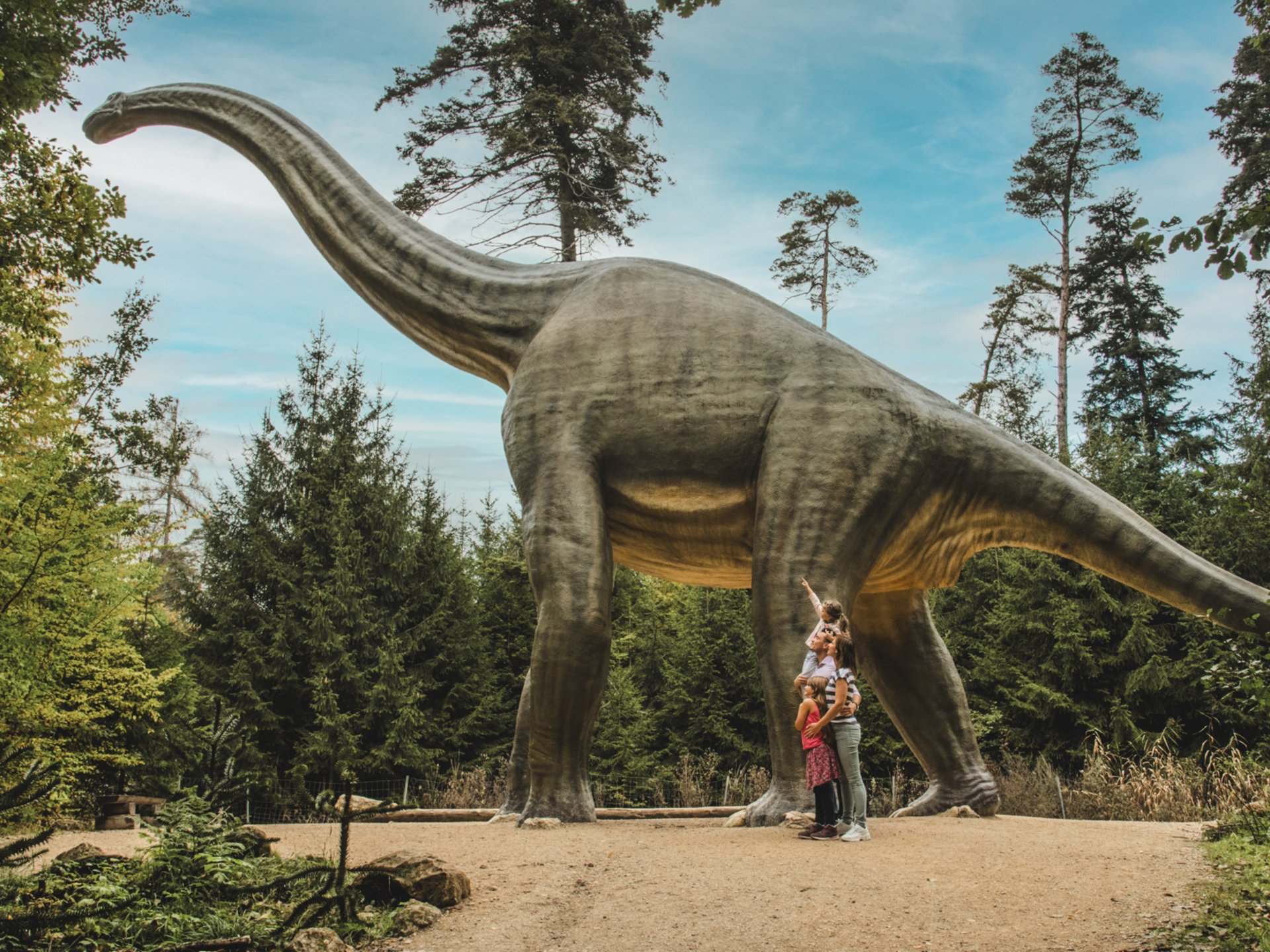 Wandern, Radfahren & Natur erleben Große Dinosaurierstatue in einem Wald mit drei Menschen darunter