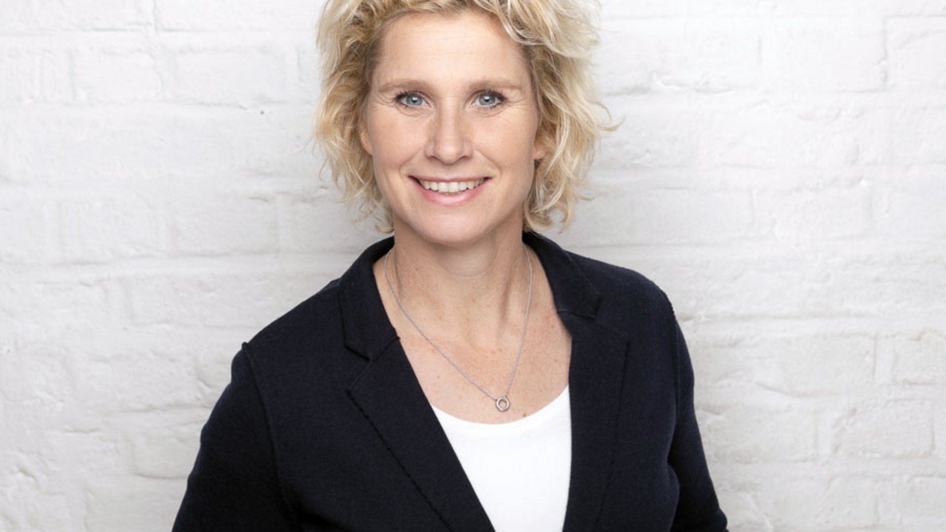 Mental Leadership Retreat Frau mit kurzen blonden Haaren lächelnd vor weißer Ziegelwand