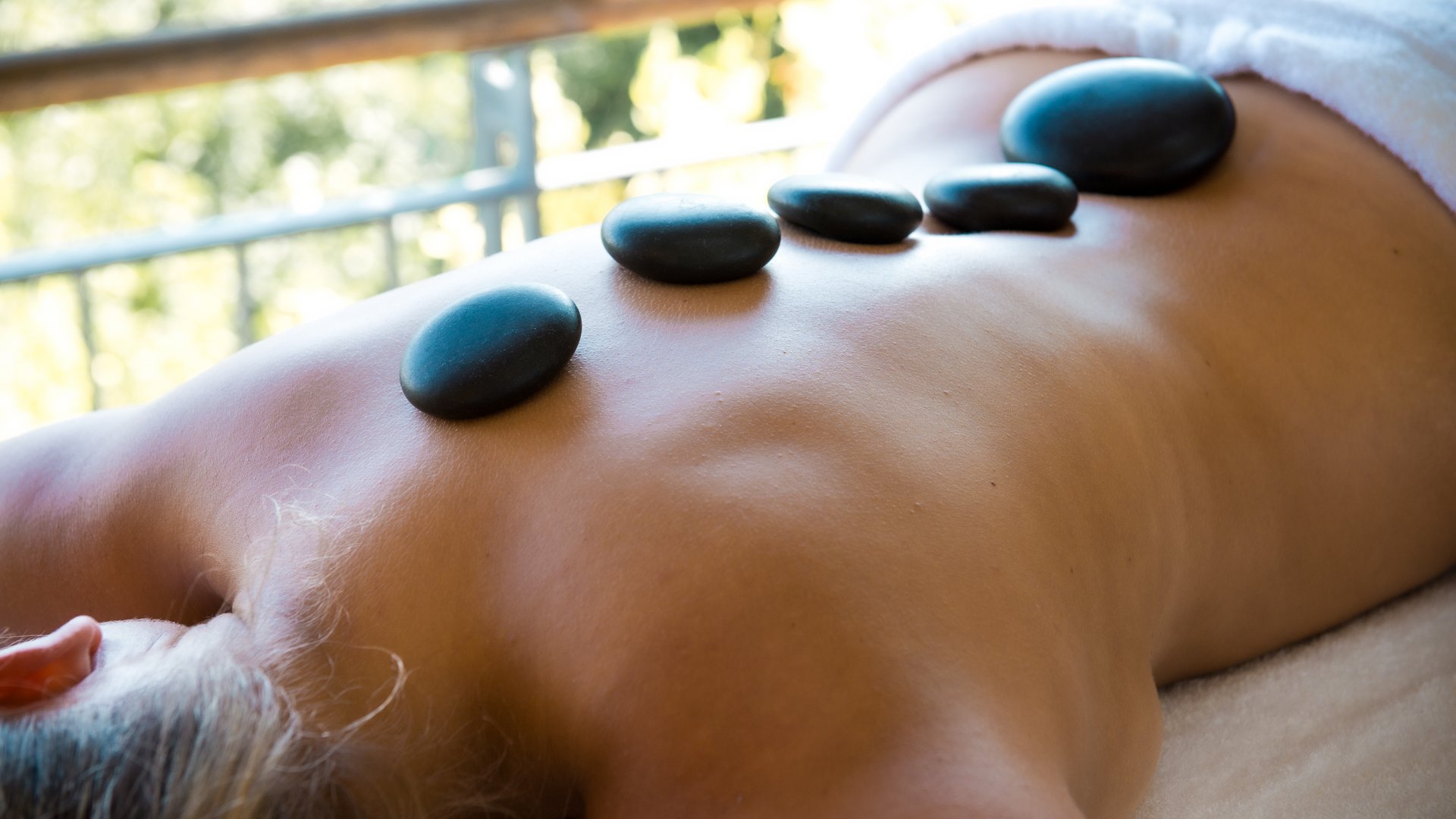 Wellness in der Oberpfalz: Massage & Beauty Person liegt mit heißen Steinen auf dem Rücken in einer Massageumgebung