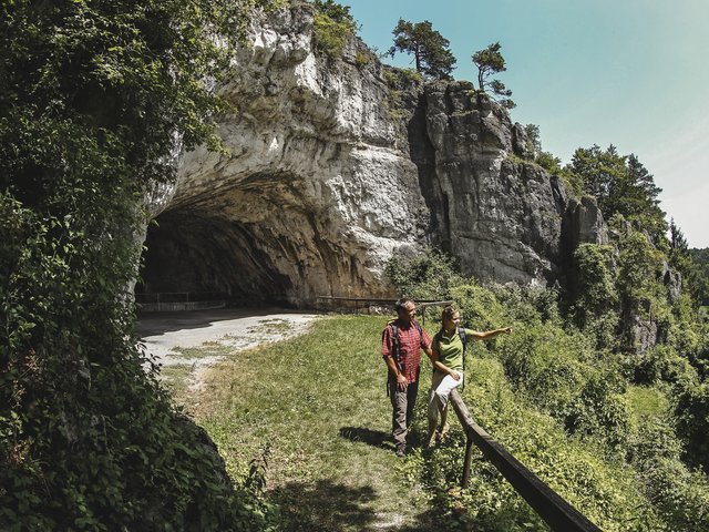 Romantik Hotel Hirschen: Bildergalerie Zwei Wanderer vor einer Felswand und Höhle in einer grünen Landschaft