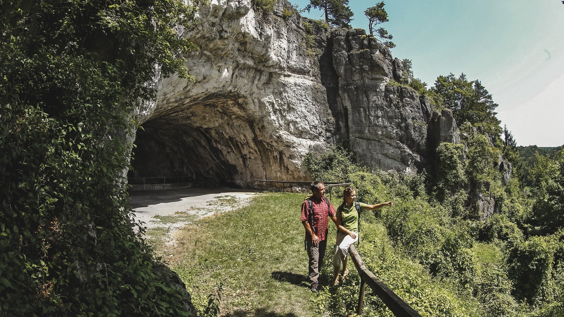 Wandern in der Oberpfalz: Aktiv in der Natur Zwei Wanderer vor einer Felswand und Höhle in einer grünen Landschaft