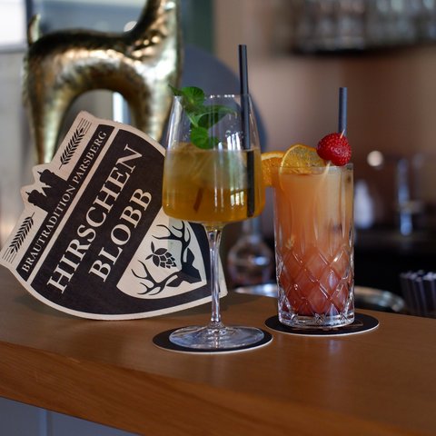 Hirschenblobb: unsere Bar Zwei Cocktails und eine Hirschfigur mit Hirschen Blobb Schild auf einer Bar