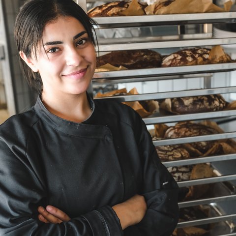 Nachhaltiges Hotel in Bayern mit eigener Manufaktur Lächelnde Bäckerin in schwarzer Arbeitskleidung vor Backblechen mit Brot
