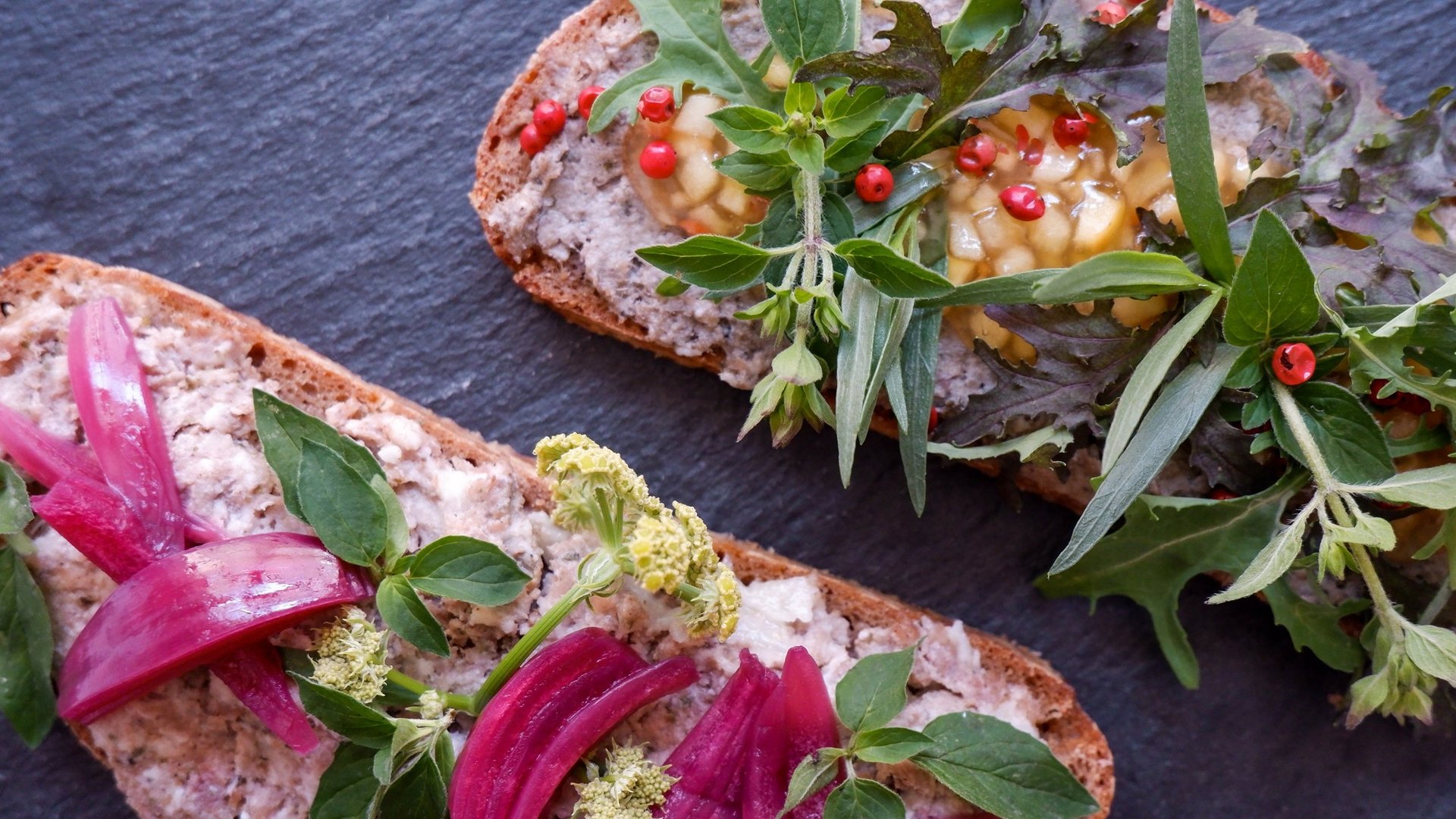 Nachhaltiges Hotel in Bayern mit Hirschenmanufaktur Belegtes Brot mit Kräutern, roten Zwiebeln und Pfefferkörnern auf dunkler Unterlage