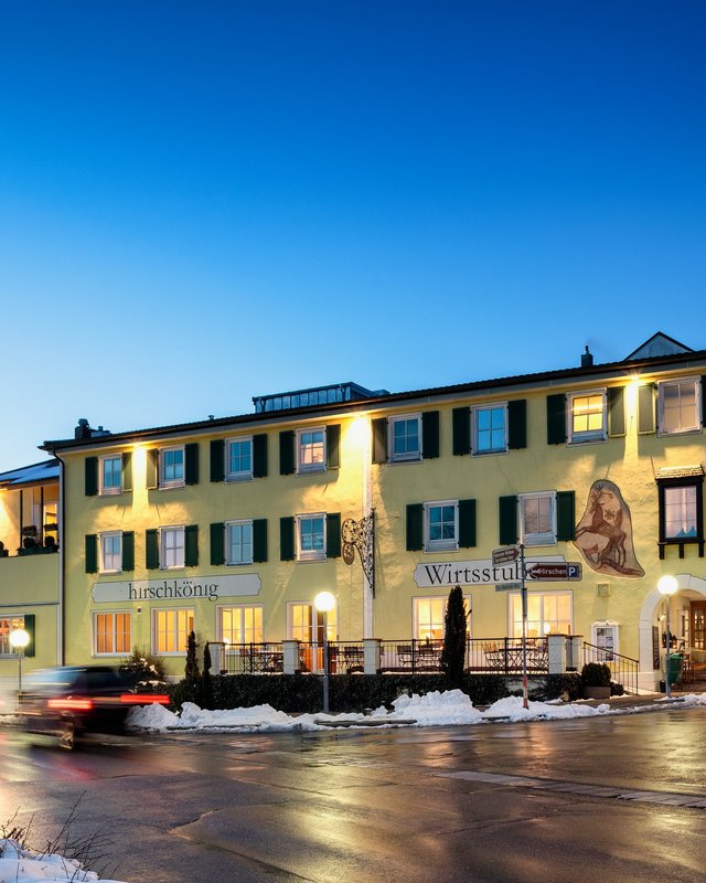 Nachhaltiges Hotel in Bayern mit Hirschenmanufaktur Beleuchtetes gelbes Gebäude bei Abenddämmerung mit Schnee und Autos auf Straße