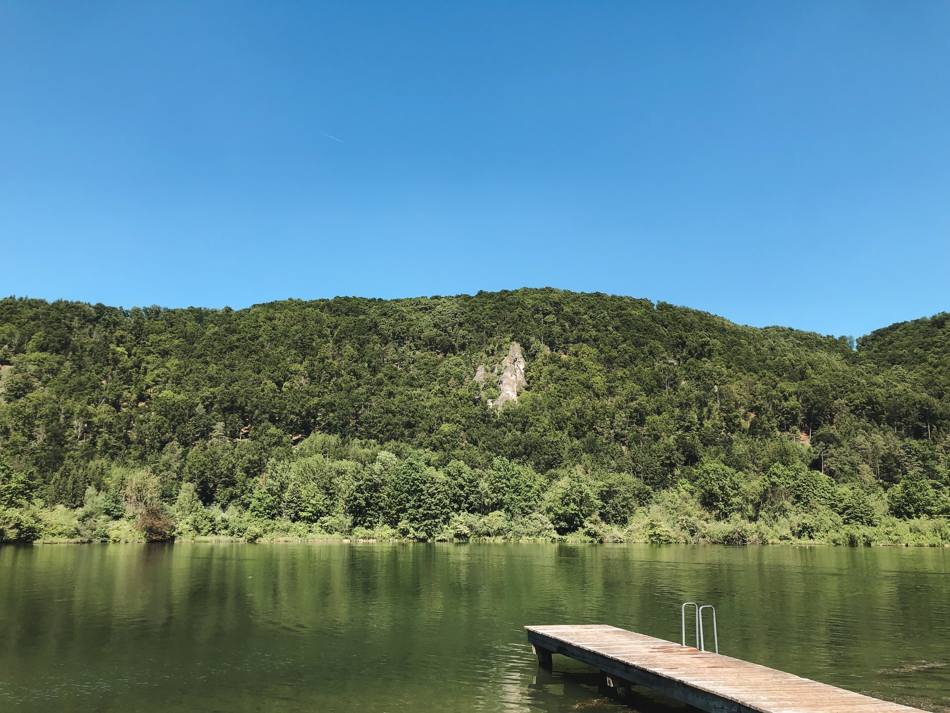 Badeseen Holzsteg am ruhigen See mit bewaldetem Hügel und blauem Himmel