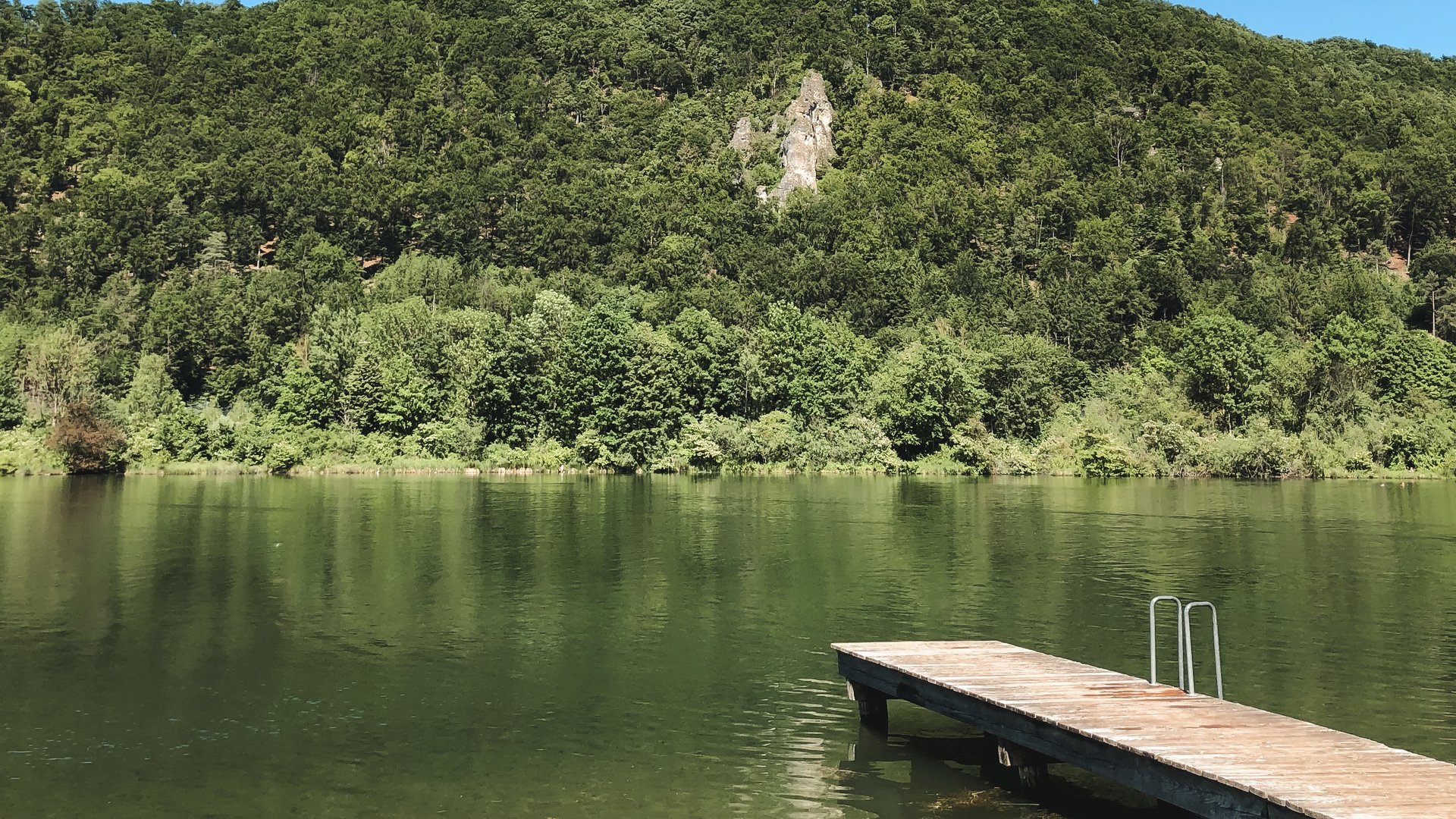 Badeseen Holzsteg am ruhigen See mit bewaldetem Hügel und blauem Himmel