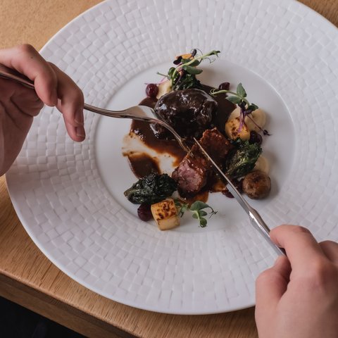 Romantik Hotel Hirschen: Gastgeber & Werte Person schneidet elegantes Gericht mit Fleisch und Gemüse auf weißem Teller