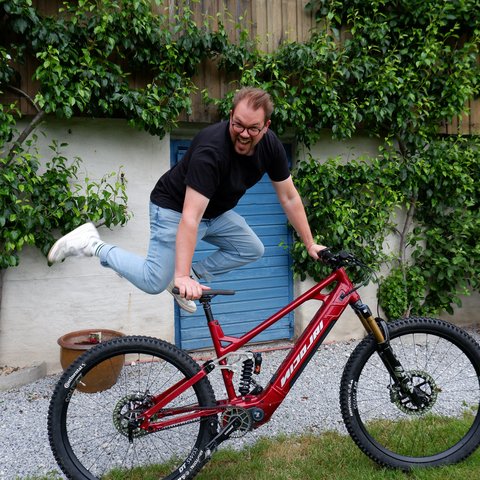 Hotel mit Fahrradverleih in Bayern gesucht? Mann springt über rotes Mountainbike vor grünem Busch und blauem Türhintergrund