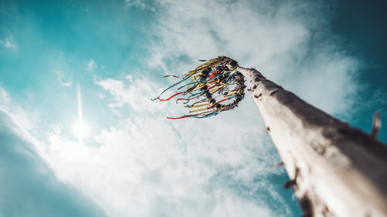Blick nach oben auf einen Maibaum mit bunten Bändern bei bewölktem Himmel