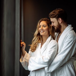 Home Paar in weißen Bademänteln schaut und lächelt aus dem Fenster