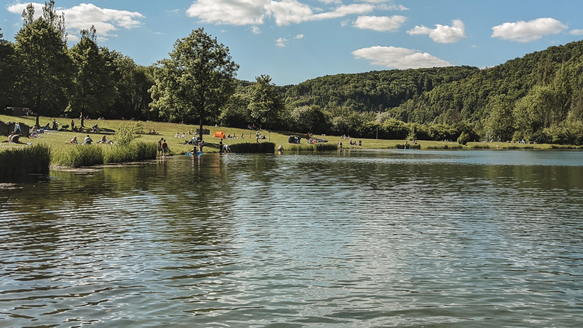 Badeseen See mit Menschen am Ufer und bewaldeten Hügeln unter blauem Himmel
