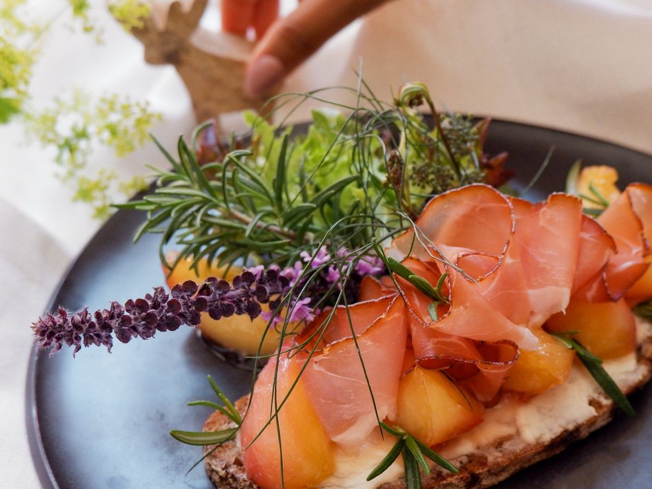 Gasthof Parsberg: Ein Gruß an den guten Geschmack Belegtes Brot mit Schinken, Melone und frischen Kräutern auf schwarzem Teller