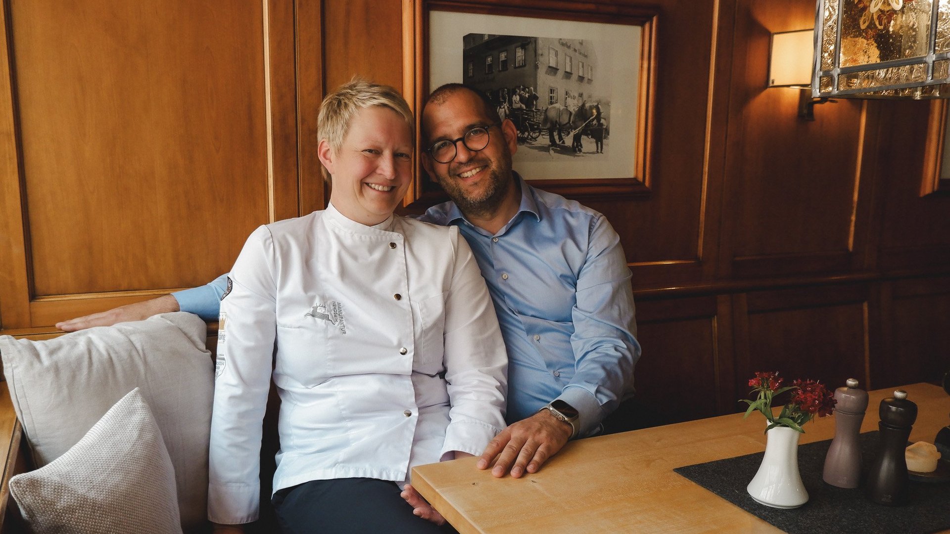 Home Two smiling people sitting at a wooden table in a cozy restaurant