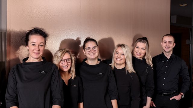 Romantik Hotel Hirschen: Blog Sechs lächelnde Mitarbeitende in schwarzer Berufsbekleidung vor beiger Wand