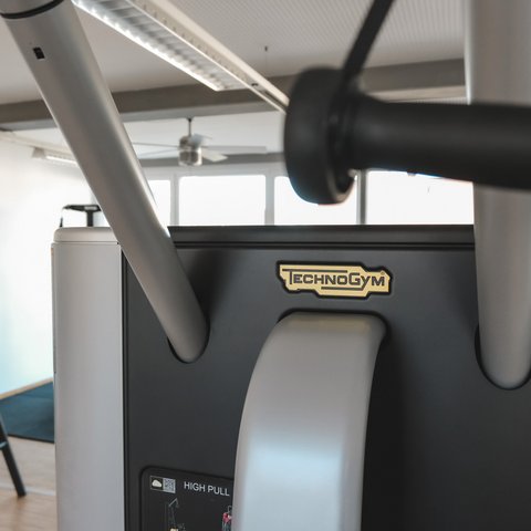 Fitnessraum Technogym fitness machine in gym with dumbbells in background