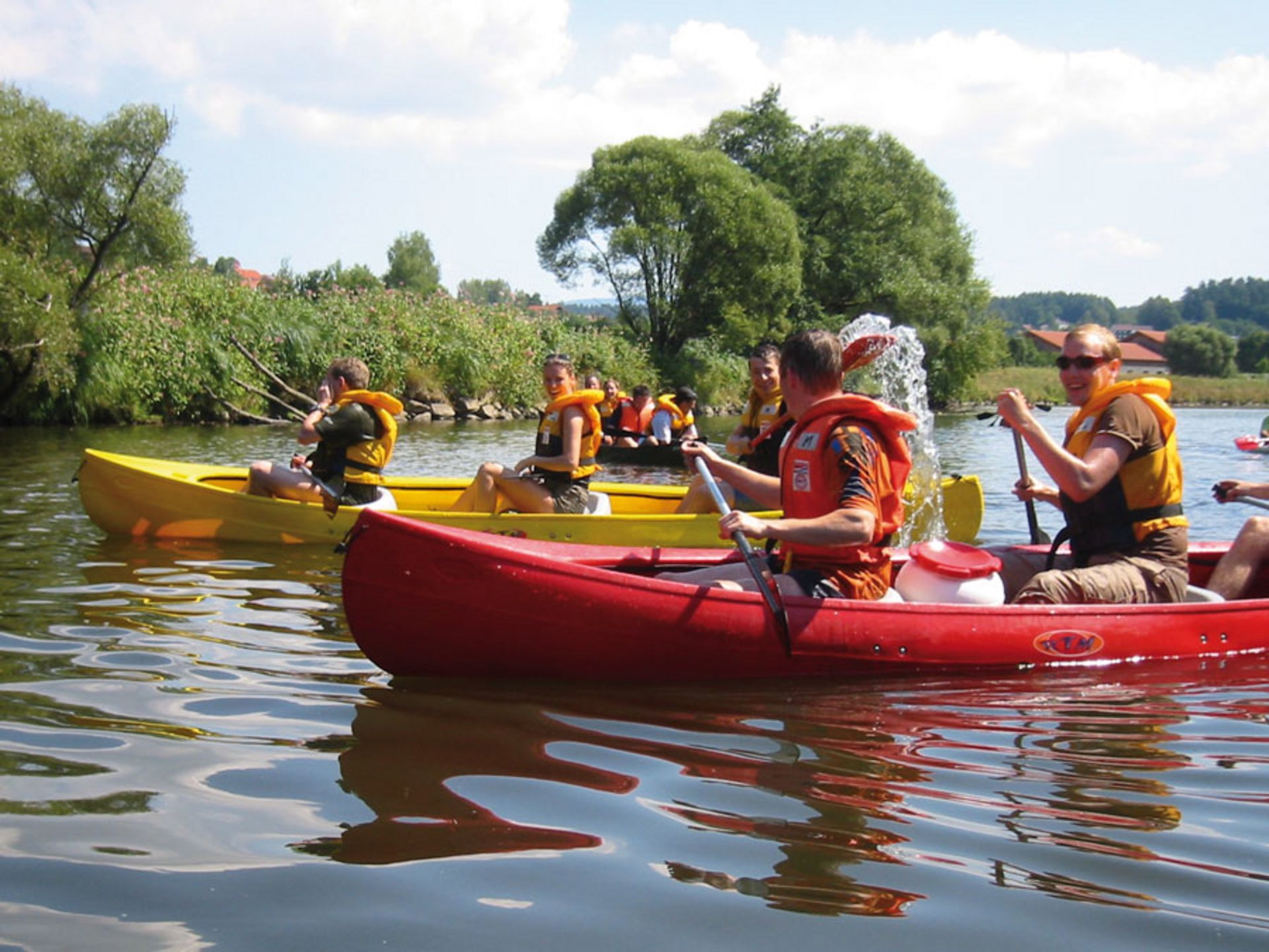 Teambuilding, Firmenevents & Rahmenprogramme Gruppe von Menschen in Kajaks auf ruhigem Fluss mit Rettungswesten bei sonnigem Wetter