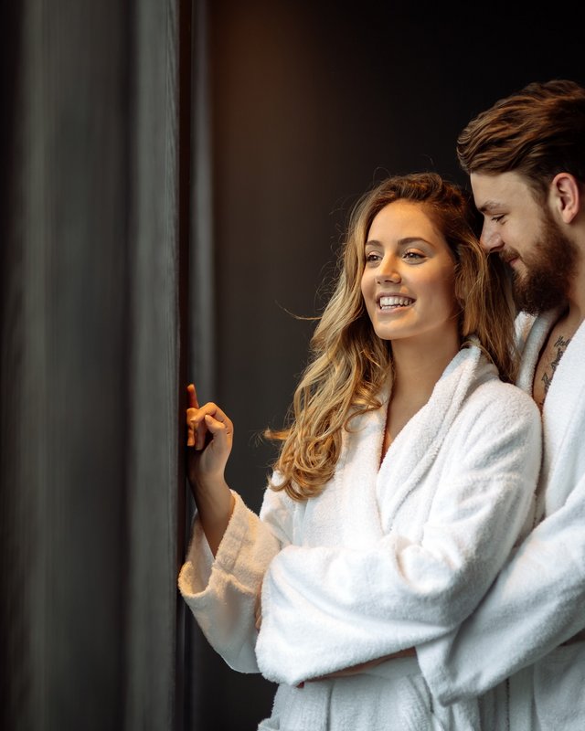 Wellnesshotel in der Oberpfalz: schwitzen & schwimmen Paar im weißen Bademantel, steht lächelnd am Fenster