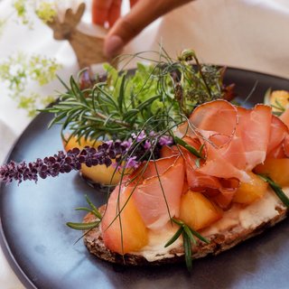 Angebote aus dem Romantik Hotel Hirschen Belegtes Brot mit Schinken, Melone und frischen Kräutern auf schwarzem Teller
