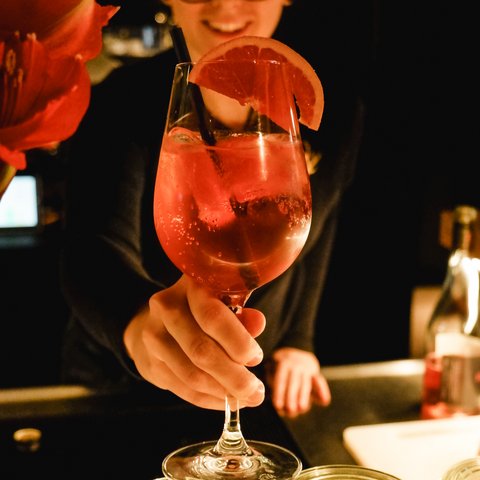 Gasthof Parsberg: Ein Gruß an den guten Geschmack Person reicht ein Glas mit rotem Cocktail und Grapefruitzeste an einer Bar