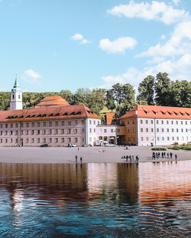 Golfurlaub Bayern: Platzhirsch oder Anfänger? Kloster mit rotem Dach am Flussufer mit bewaldeten Klippen im Hintergrund