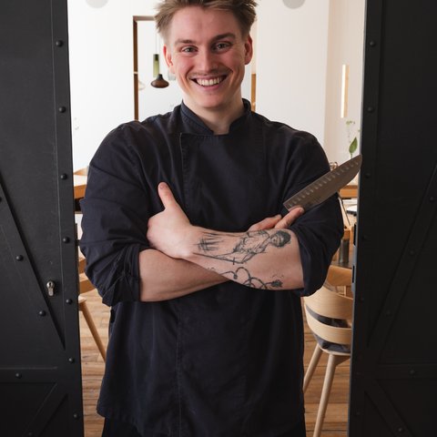 Restaurant in Parsberg: Gourmet genießen Junger Koch in schwarzer Kleidung mit Messer im Arm, lächelnd