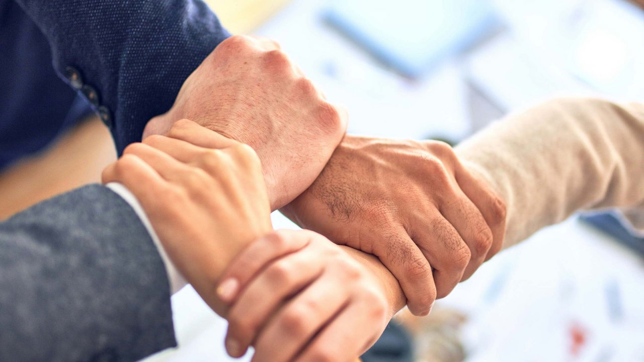 Vier Hände greifen sich in einem Teammeeting zum symbolischen Zusammenhalt